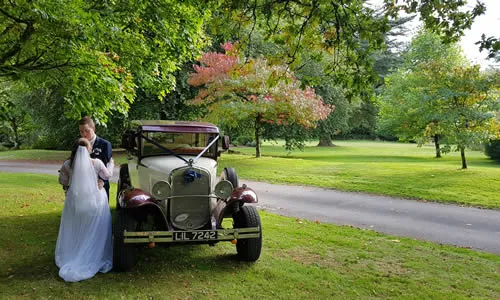 A vintage Beauford convertible parked beside a stone garden wall, white ribbons fitted.