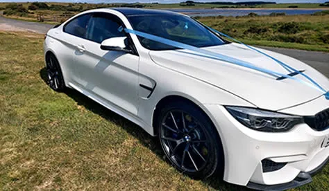 BMW M4 in Alpine white with blue ribbons fitted, parked on a coastal headland.