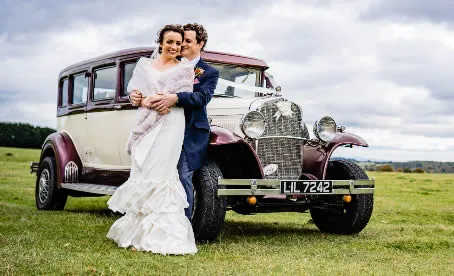 A bride and groom embracing in front of a vintage Bramwith with old English white body and burgundy fenders.