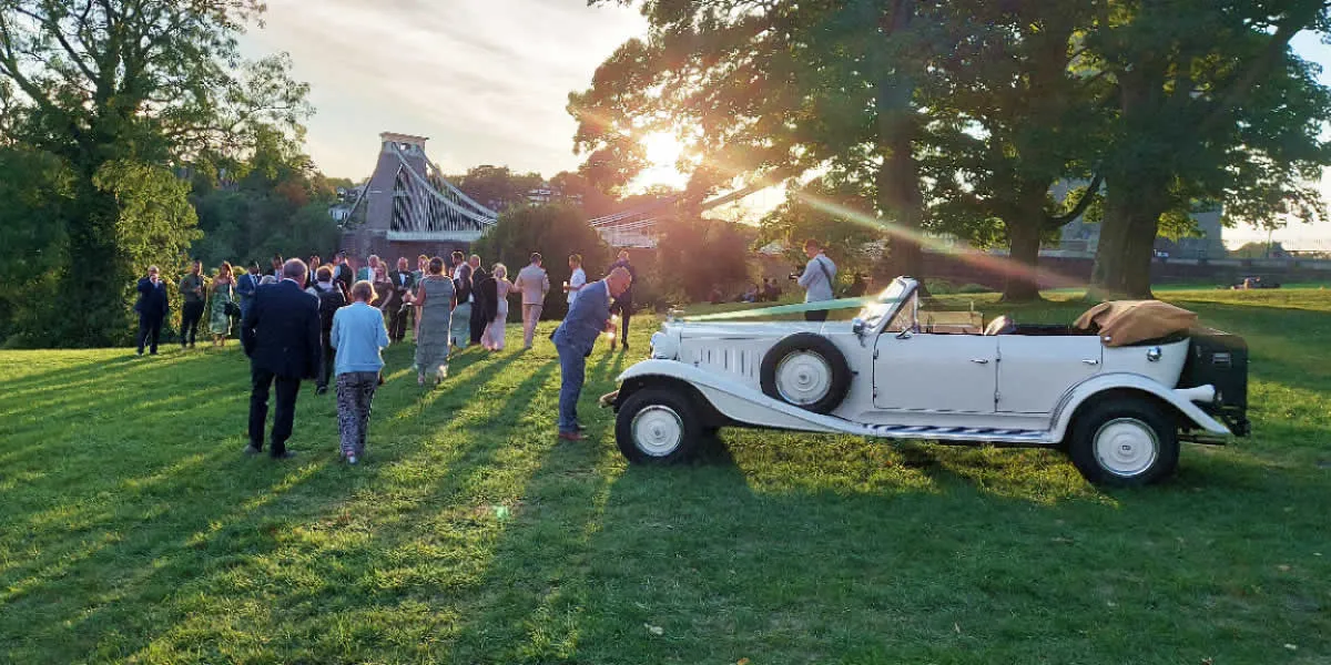The Beauford convertible at Clifton Suspension Bridge as the sun sets through the trees — wedding party walking towards the bridge.