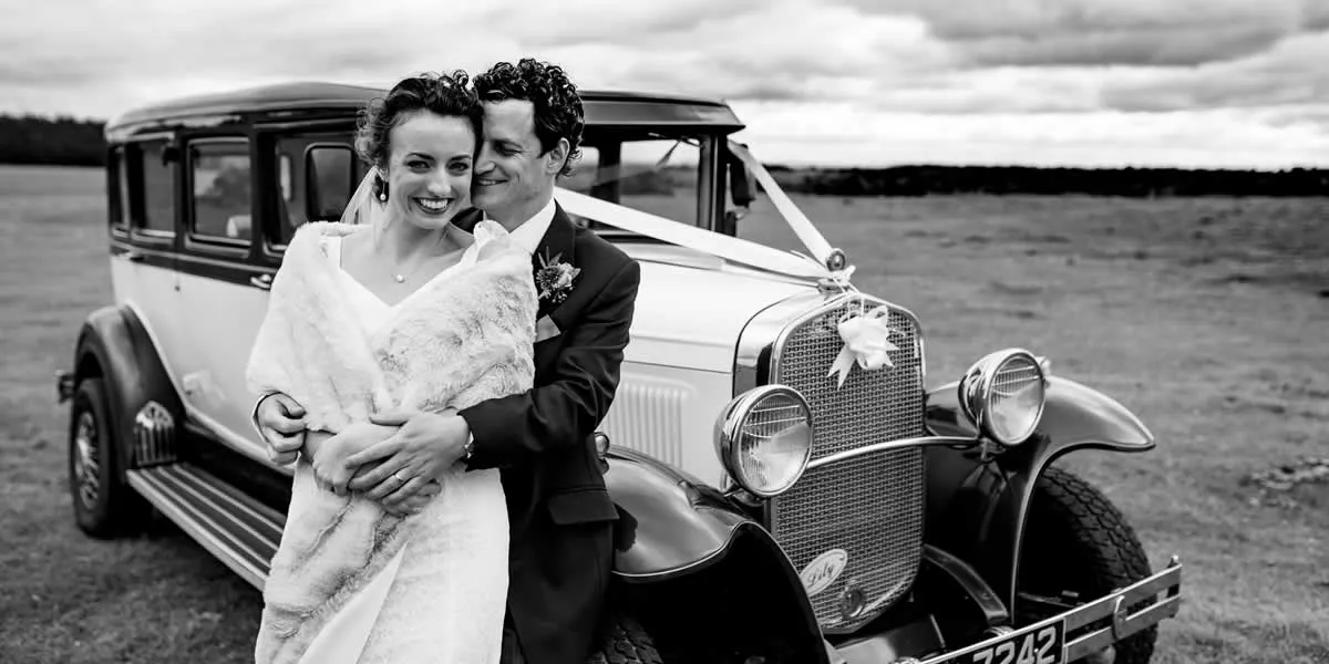 A bride and groom in wedding attire embracing in front of a vintage 1929 Bramwith on the open moorland — black-and-white photograph.